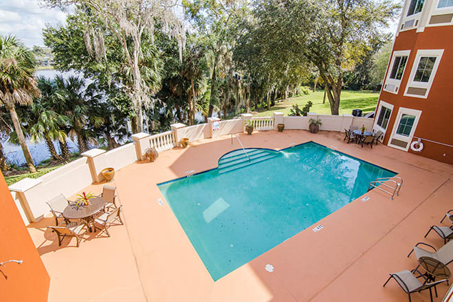 A tranquil outdoor swimming pool area with patio seating surrounded by lush greenery on a sunny day.
