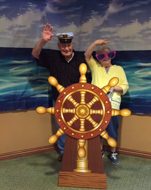 A playful photo with two people posing behind a ship's wheel, wearing a captain's hat and oversized glasses, against a nautical backdrop.