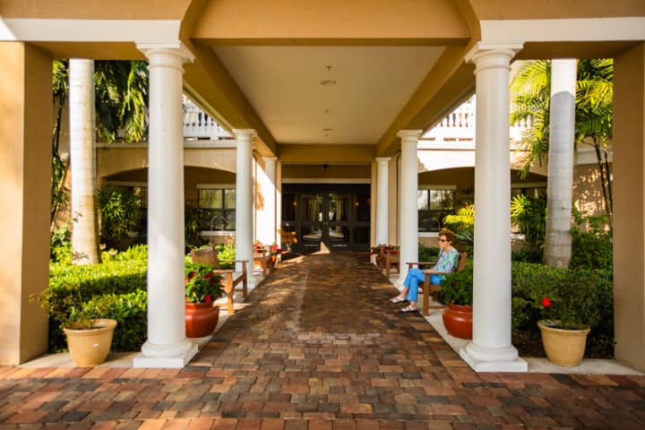 A covered passageway with columns and a brick walkway, featuring outdoor seating and surrounding garden plants.