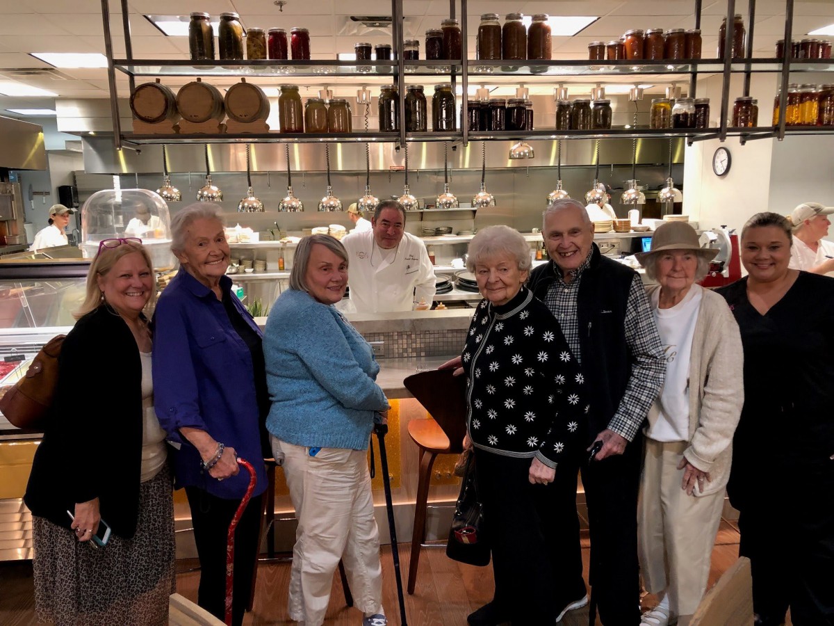 A group of elderly individuals standing in front of a restaurant kitchen with a smiling chef in the background.