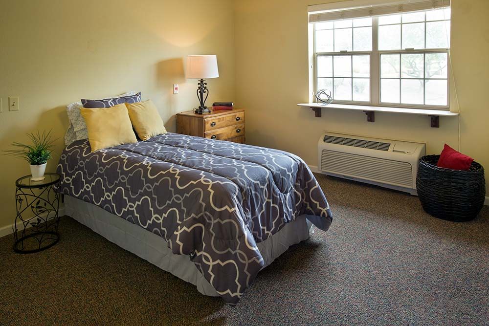 A cozy bedroom with a neatly made bed, bedside table, and a window letting in natural light.