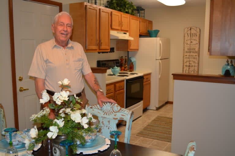 A man standing in a well-decorated kitchen with wooden cabinets and a dining area featuring a floral centerpiece.