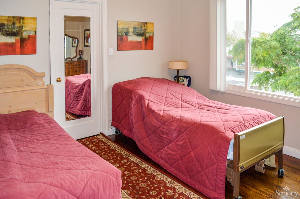 A cozy bedroom with twin beds covered in red bedding, featuring a mirror, lamp, and a window with natural light.