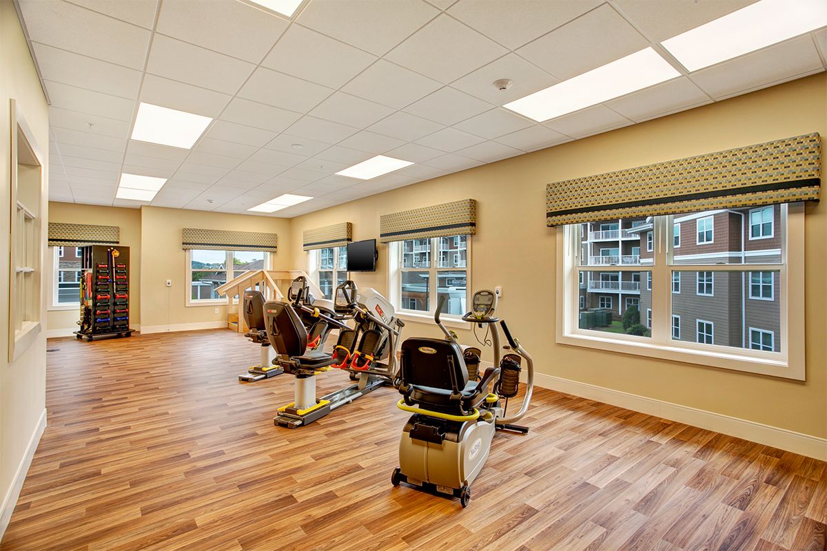 A bright and spacious fitness room with exercise equipment, including treadmills and stationary bikes, set against large windows allowing natural light.