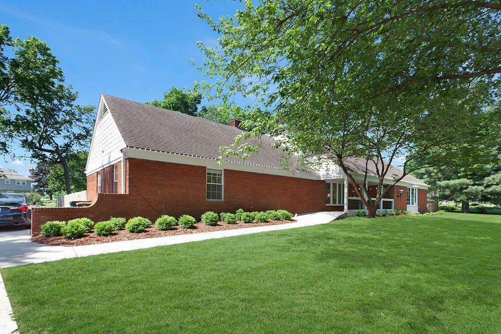 A suburban brick house with a well-maintained lawn and a tree in the yard.