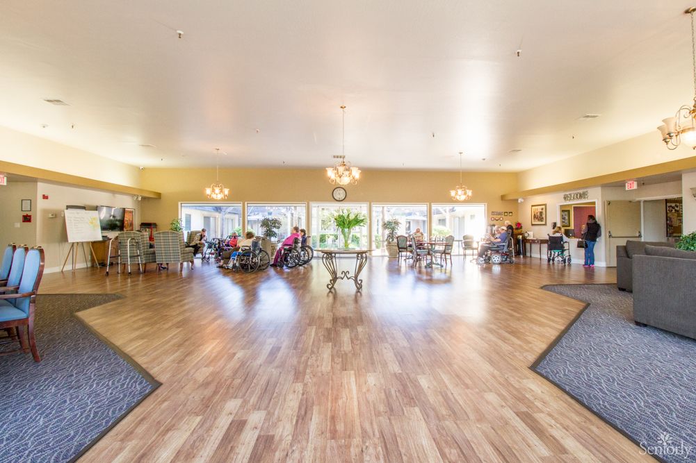 A spacious and bright community room in a senior living facility, featuring wooden flooring and elegant decor.
