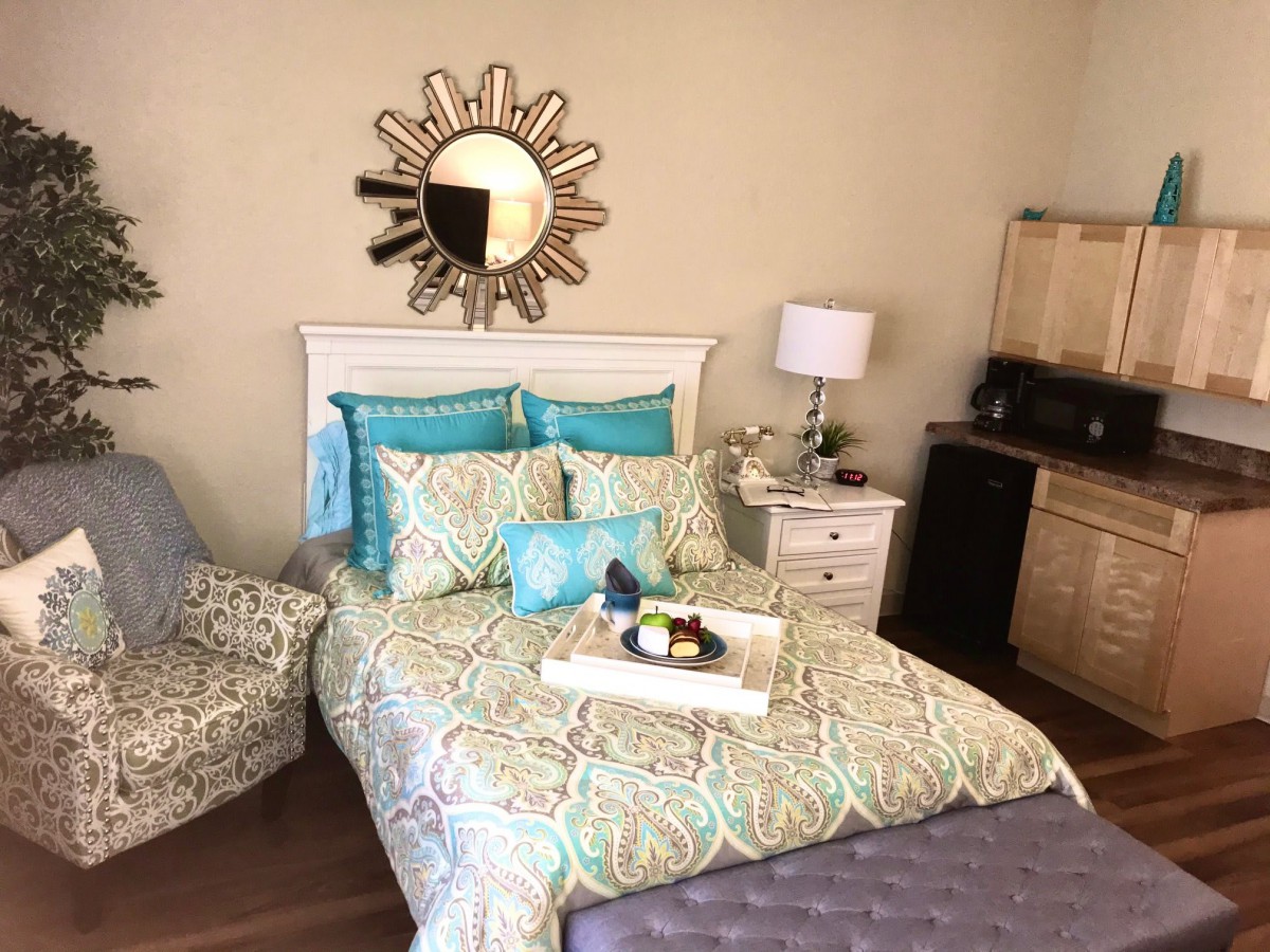 A cozy bedroom with vibrant turquoise and beige bedding, a decorative sunburst mirror above the bed, and a stylish side table with a lamp and a wooden desk.