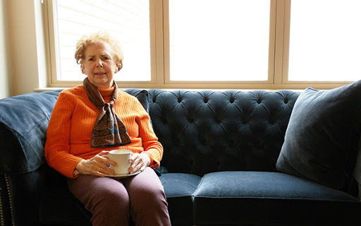 An elderly woman sits comfortably on a tufted sofa, enjoying the warm natural light coming through the windows.