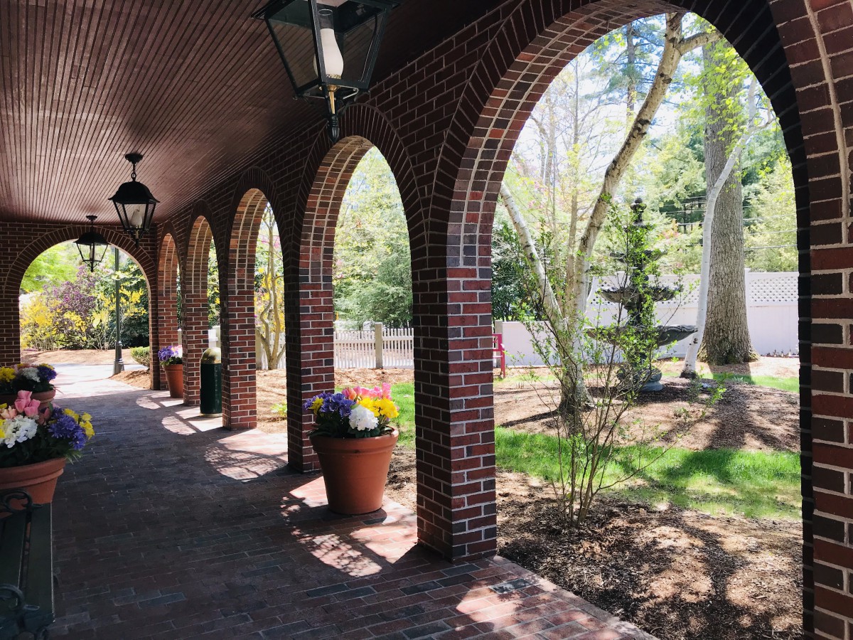 A brick archway pathway with potted plants leads to a sunlit garden.