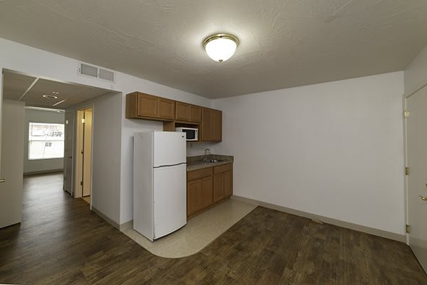 A small apartment kitchen with wooden cabinets, a white refrigerator and microwave, and hardwood flooring.