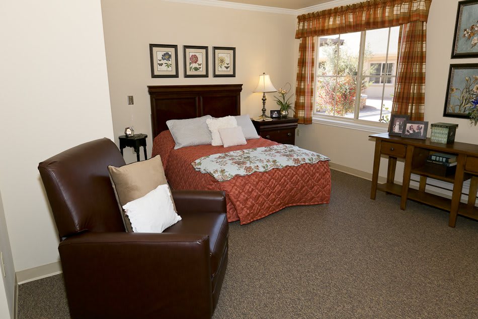 A cozy bedroom with a bed, armchair, and window, featuring warm decor and soft lighting.