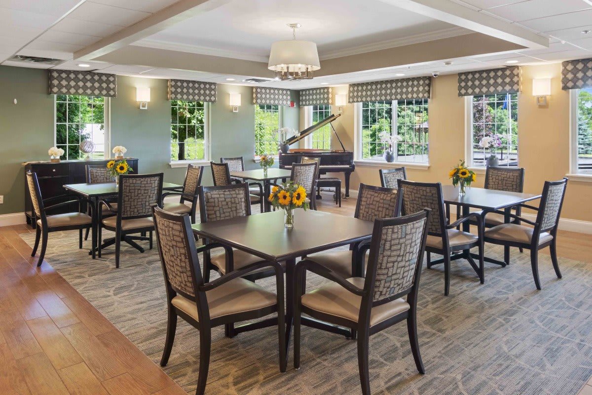 A spacious dining room with wooden tables and chairs, featuring large windows and a piano.