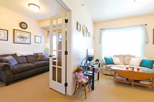 A cozy living room with a sofa, coffee table, and decorative items, featuring French doors and a doll on a chair.