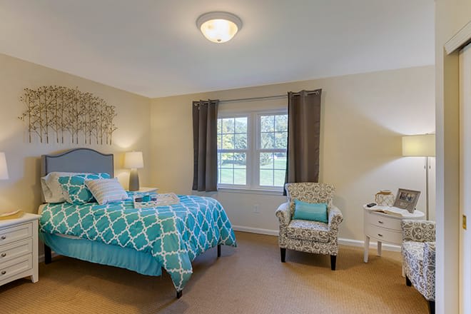 A modern bedroom featuring a teal bedspread, an armchair, and natural light from a large window.
