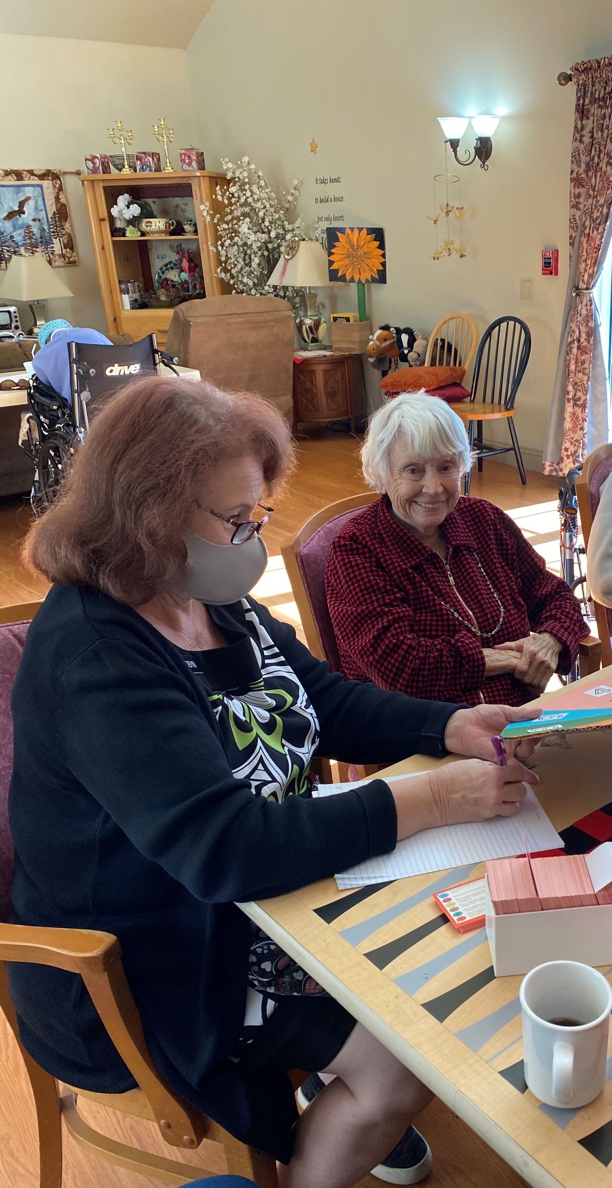 Two women sitting at a table, with one wearing a mask and engaging in an activity.