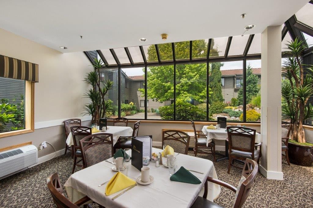A bright dining area with large windows overlooking a garden, featuring neatly set tables and chairs.