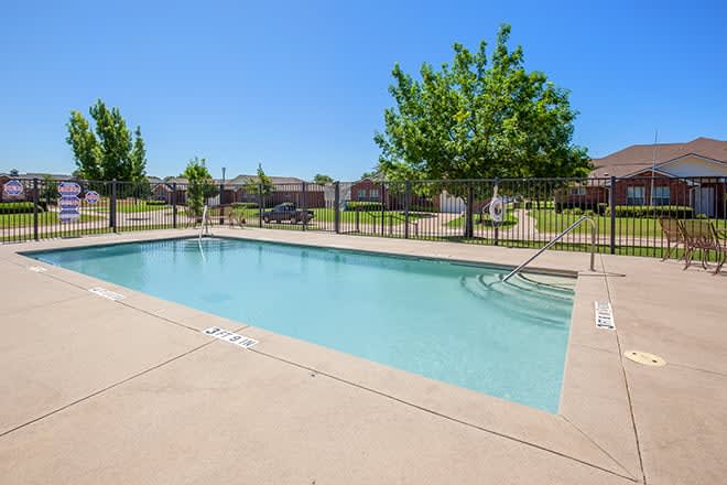 A sunny day by an outdoor swimming pool surrounded by a patio and fence, with trees and a clear blue sky in the background.