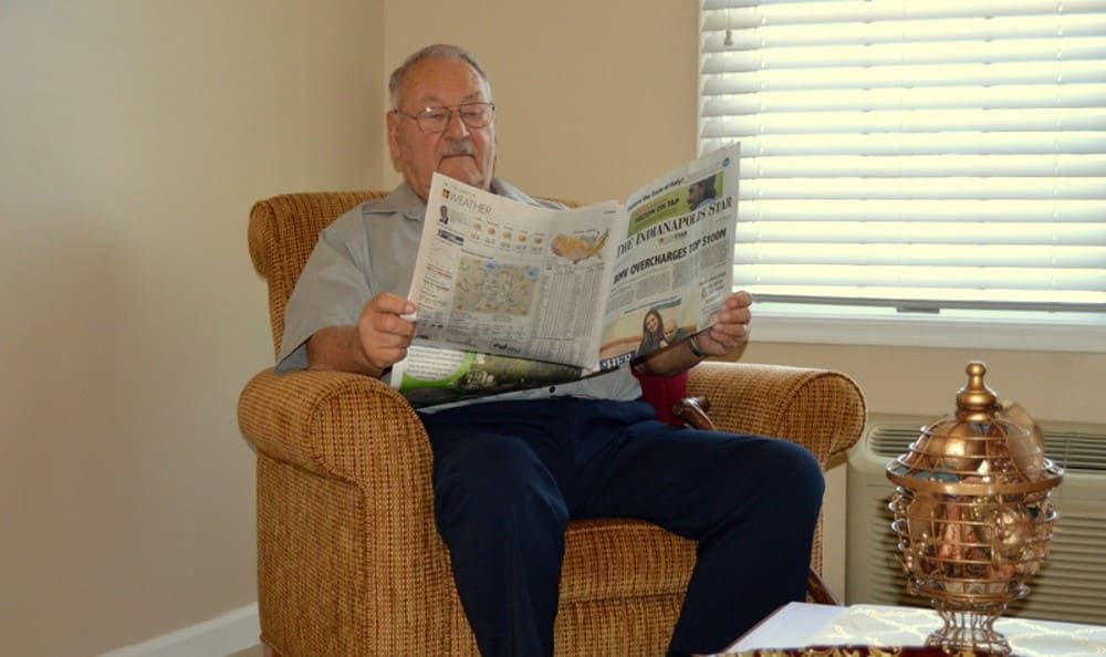 A senior man sitting in an armchair reading a newspaper indoors.