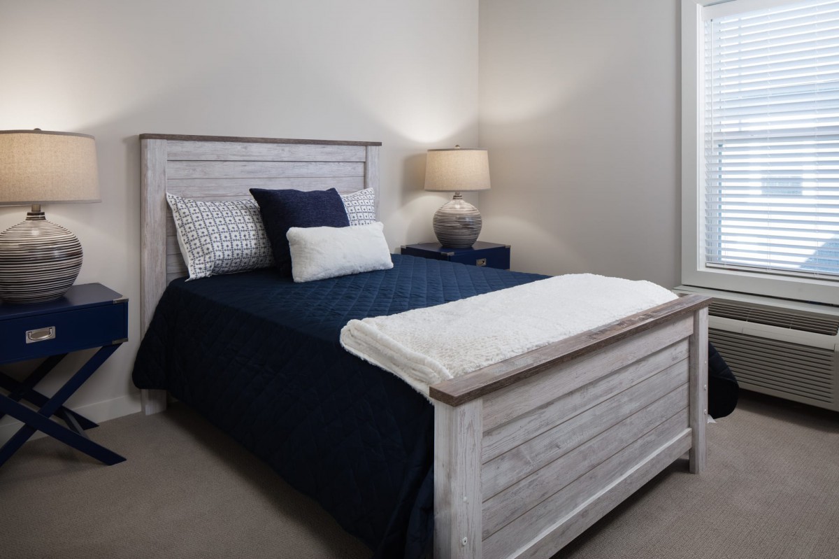 A cozy bedroom with a neatly made bed, featuring navy and white bedding, accompanied by a bedside lamp on a nightstand.