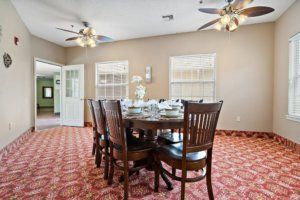 A dining room with a round wooden table set for a meal, surrounded by chairs and featuring ceiling fans and patterned carpet.