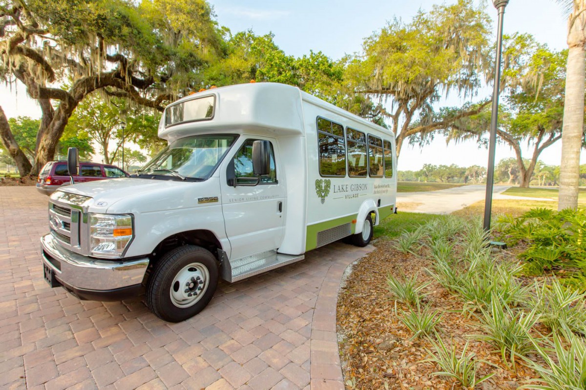 A white shuttle bus parked on a paved path, surrounded by trees and greenery.