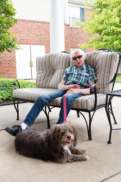 A man wearing sunglasses sits casually on an outdoor bench with a dog by his side.