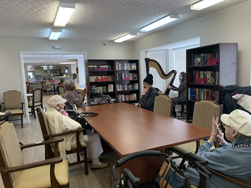 A peaceful library setting with seniors gathered around a table, surrounded by shelves of books and a harp in the corner.
