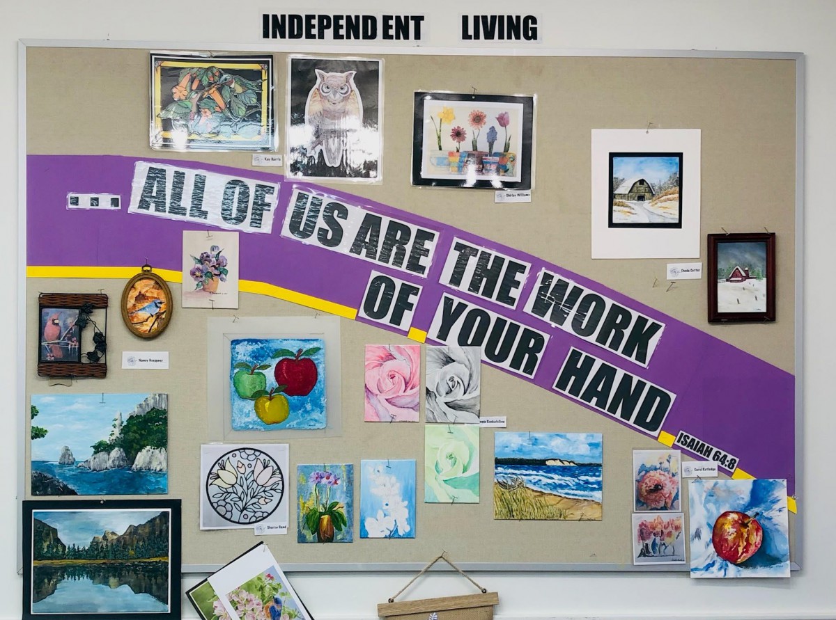 A community art display showcasing various handmade artworks with the banner 'All of Us Are the Work of Your Hand' under the theme of independent living.