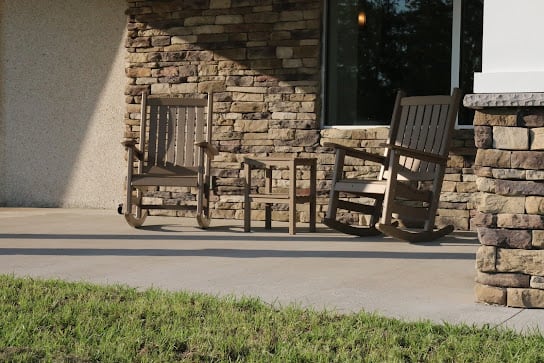 Two wooden rocking chairs on a sunny porch against a brick wall.