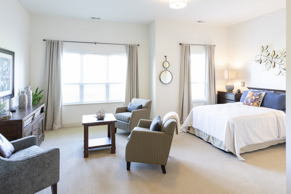 A modern, bright bedroom with large windows, featuring a bed and comfortable armchairs.