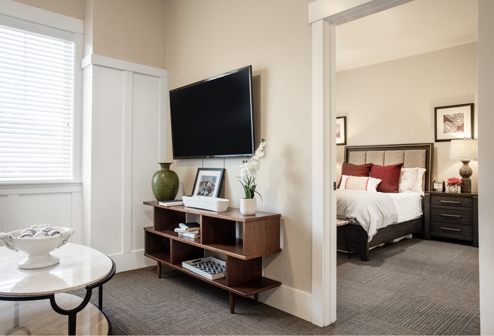 A modern interior featuring a living area with a TV, shelves, and decor leading into a cozy bedroom.