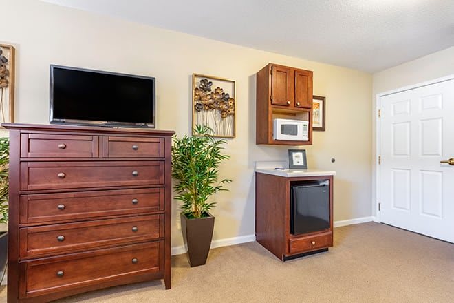 A cozy bedroom corner featuring a dresser with a TV, a microwave and mini fridge unit, and decorative indoor plants.