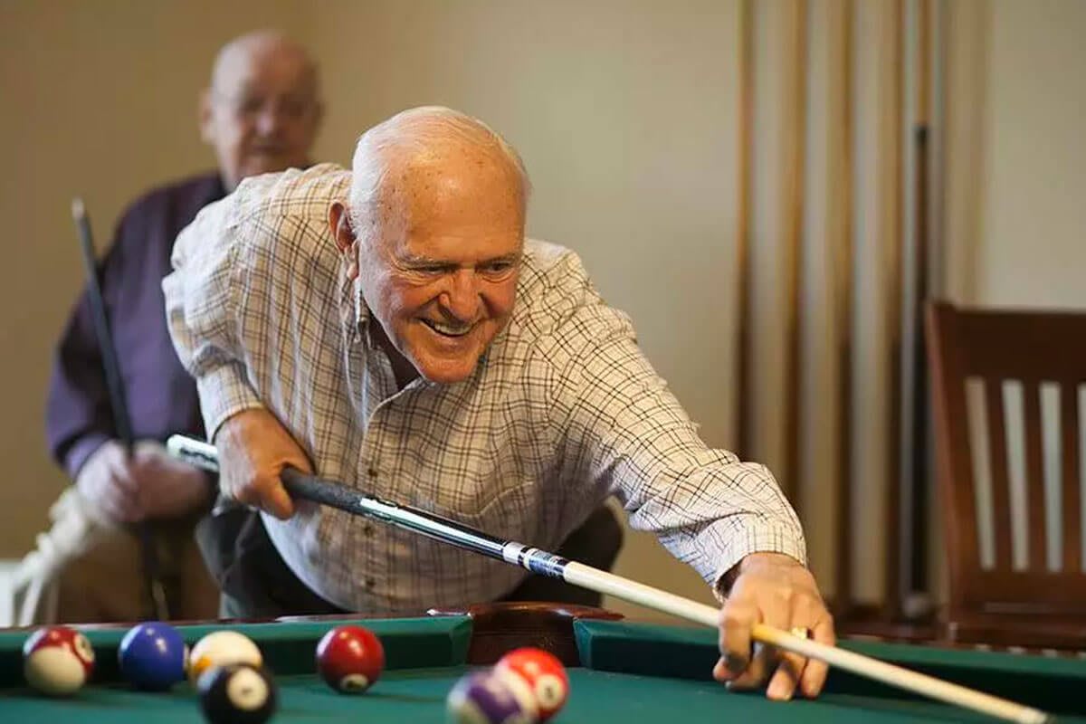 An elderly man smiling as he plays a game of pool.