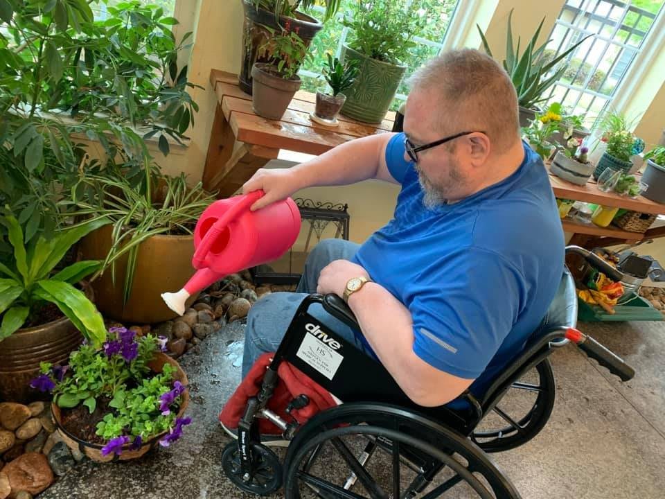 A man in a wheelchair waters indoor plants with a red watering can.