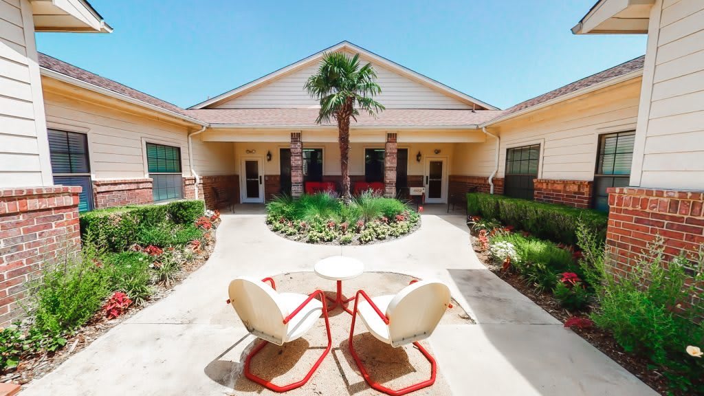 A sunny courtyard featuring a central garden with a palm tree, surrounded by buildings, and two chairs with a table in the foreground.