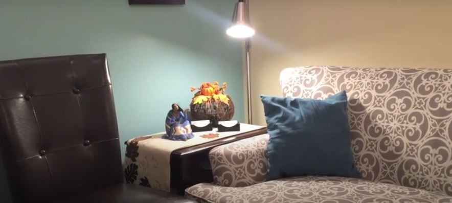 A cozy seating area featuring a patterned chair with a blue pillow, a side table with decorative items, and a floor lamp providing warm lighting.