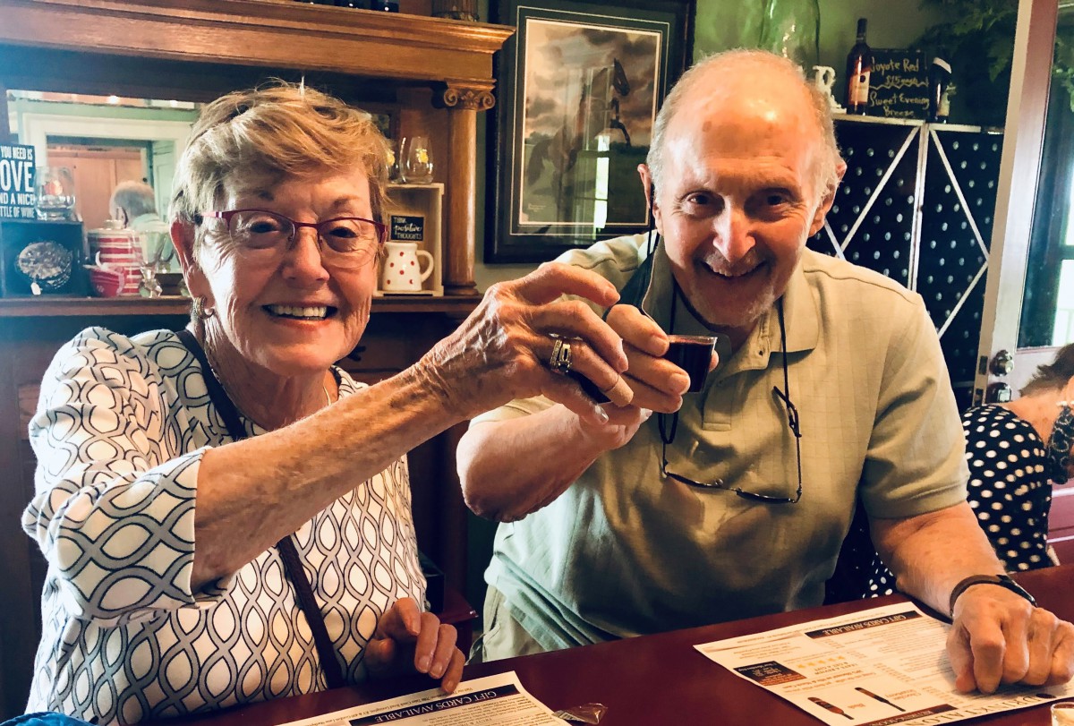 An elderly couple smiling and toasting their drinks in a cozy indoor setting.