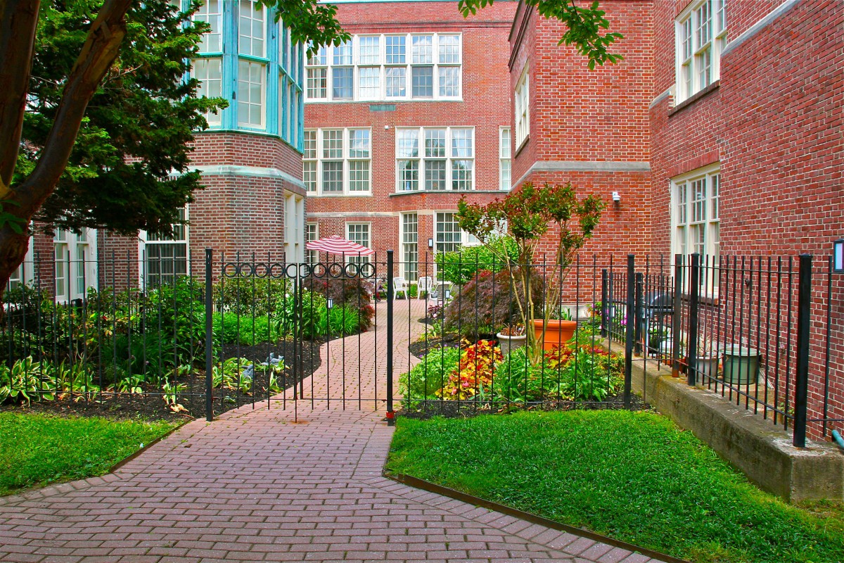 Charming garden nestled between red brick buildings with a gated pathway and lush greenery.