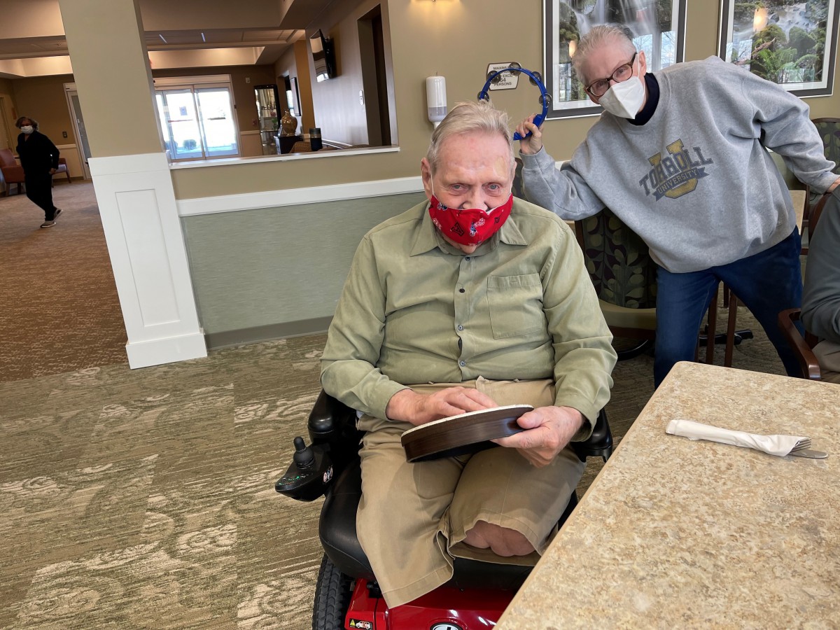 Two elderly individuals wearing masks, one seated in a wheelchair indoors.