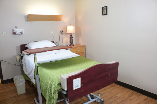 A clean and simple hospital room featuring an adjustable bed with a green blanket, a bedside lamp, and minimalistic decor.