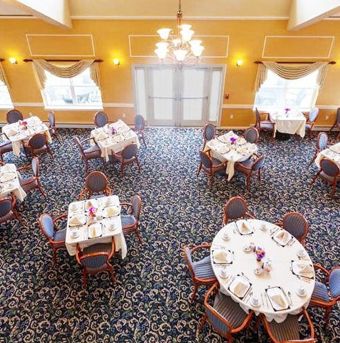 An elegant dining room with round tables and place settings, featuring a chandelier and a carpeted floor.