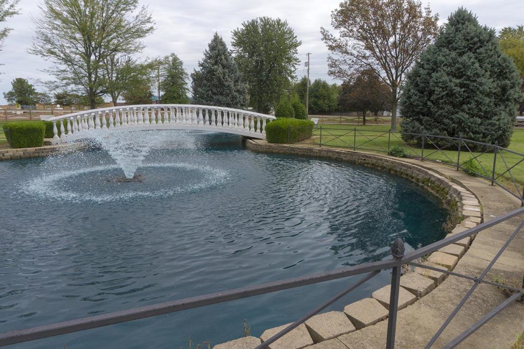 A serene park scene with a white bridge over a pond featuring a central fountain.