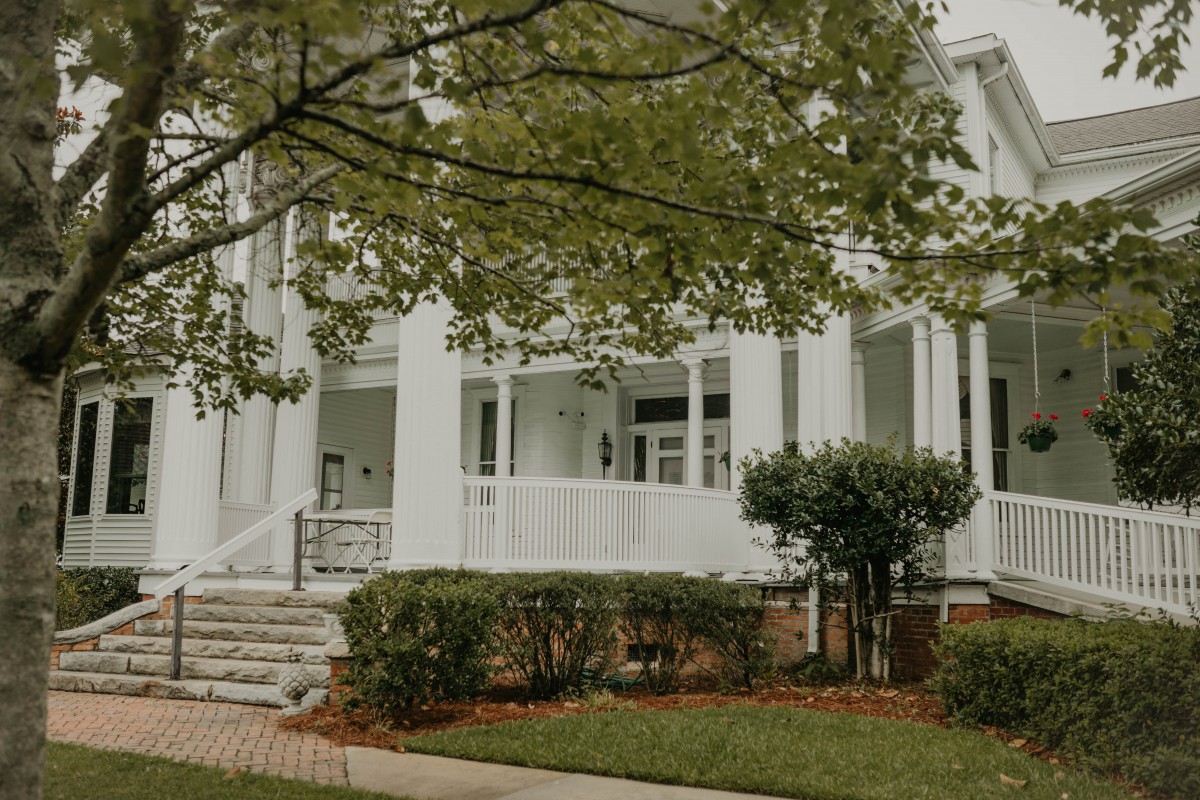 A white house with a porch, surrounded by trees and a neatly trimmed garden.