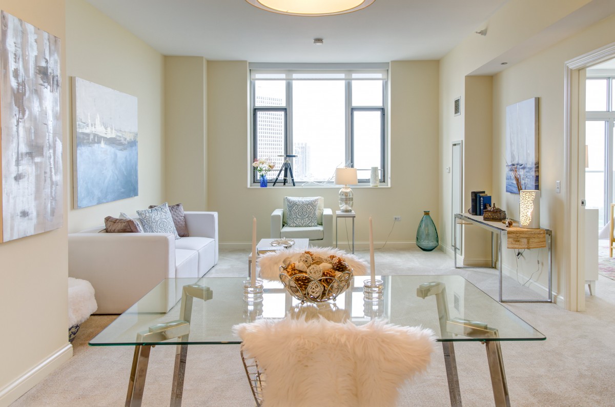 A modern and minimalistic living room with natural light streaming through the window, featuring a glass table and neutral-toned furniture.