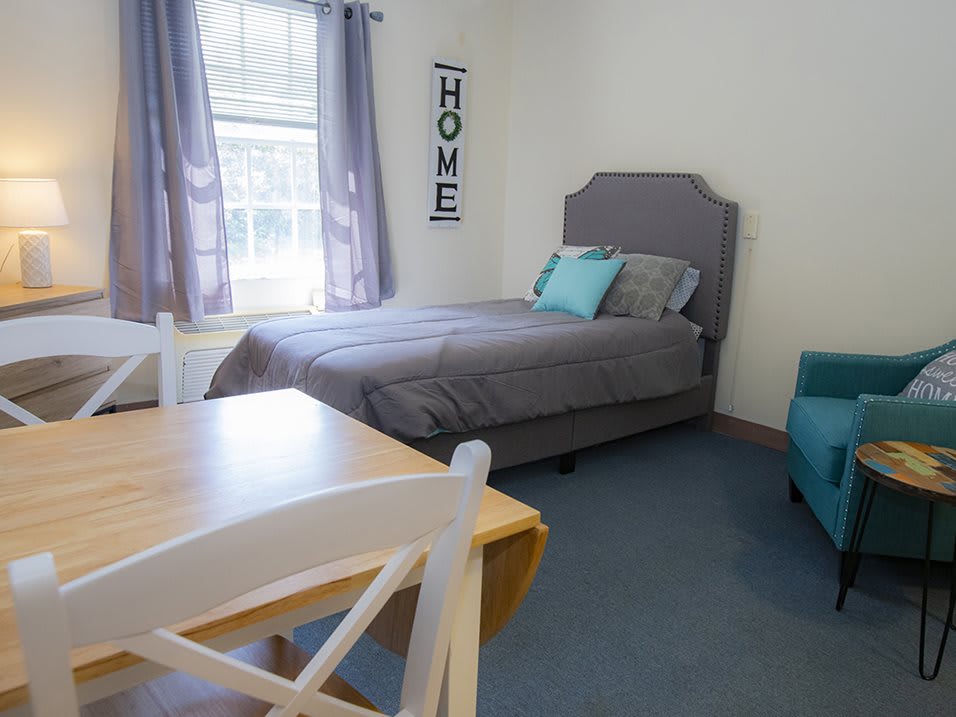 A cozy bedroom with a single gray-bedded setup, accompanied by a wooden table and chair next to a blue sofa, under soft natural light from the window.
