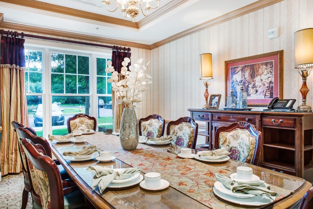 An elegantly furnished dining room featuring a well-set table with ornate chairs, a decorative vase centerpiece, and ambient lighting from a chandelier overhead.