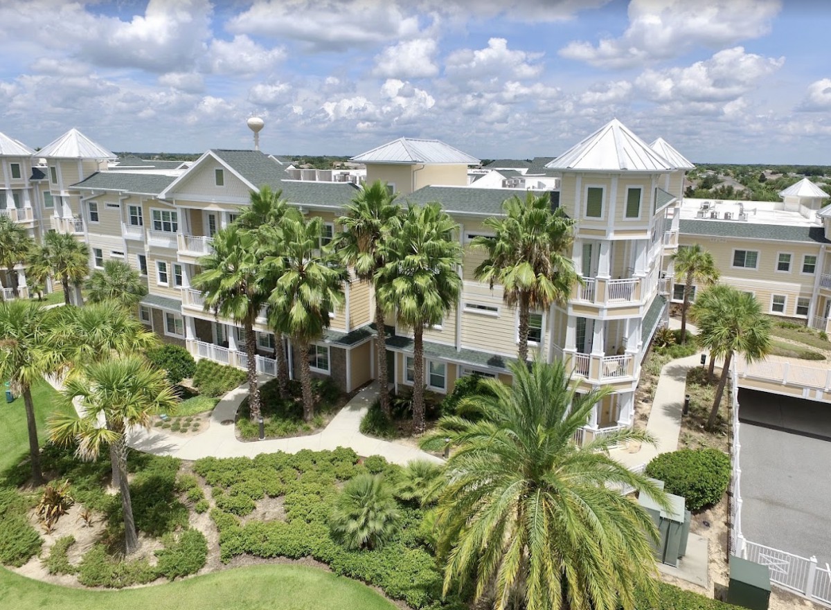 A multi-story residential building surrounded by lush greenery and palm trees under a partly cloudy sky.