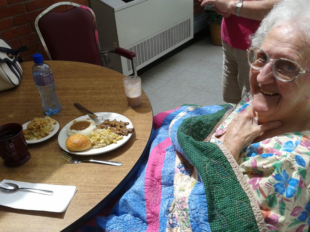 An elderly woman sitting at a dining table with a meal and wrapped in a blanket.