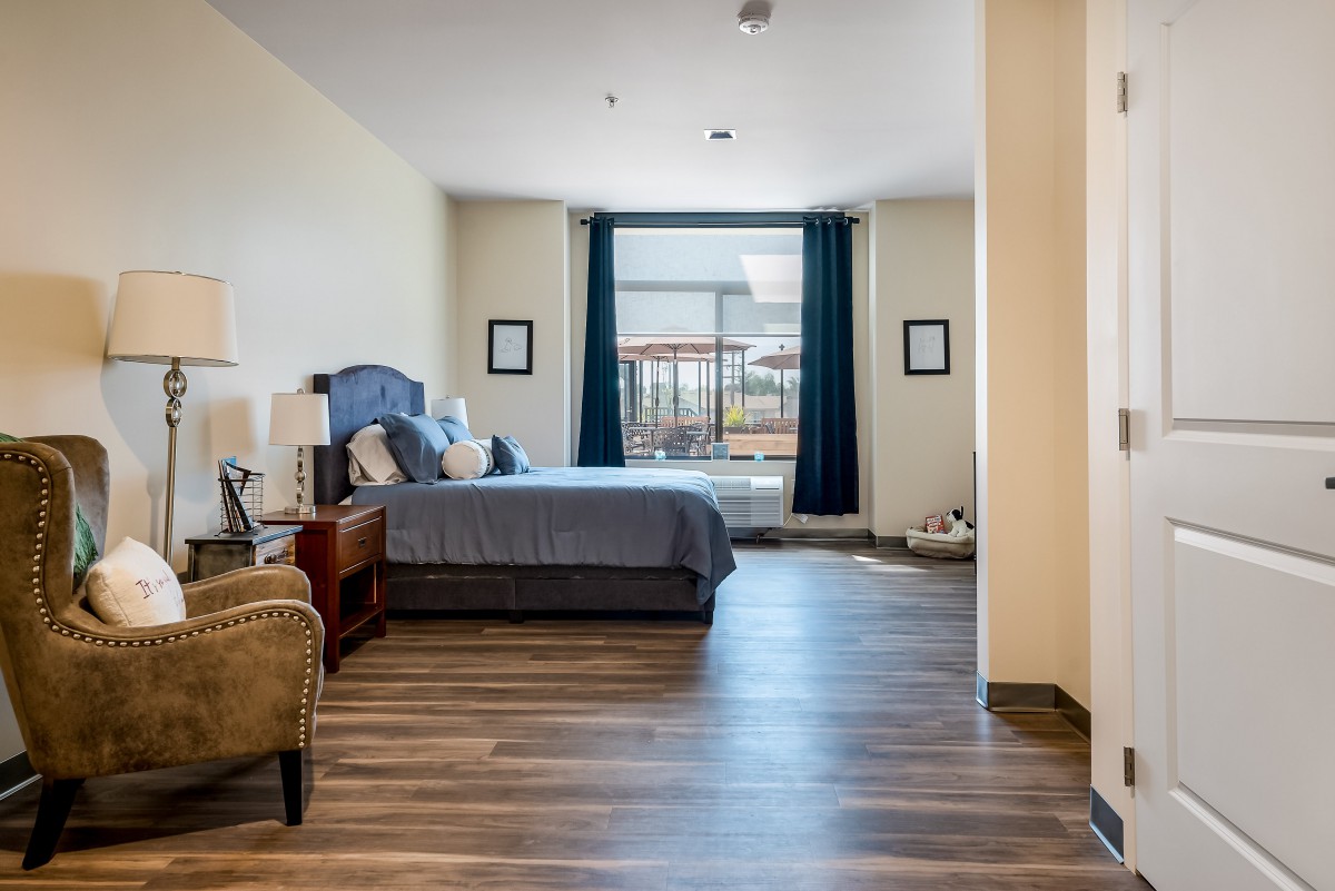 A spacious bedroom with a large bed and armchair, featuring hardwood floors and a window with curtains.