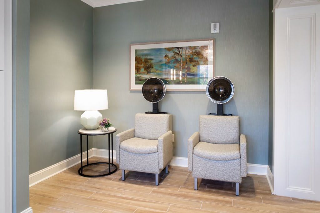 A cozy salon waiting area featuring two chairs with hair dryers, a side table with a lamp, and a tranquil painting on the wall.